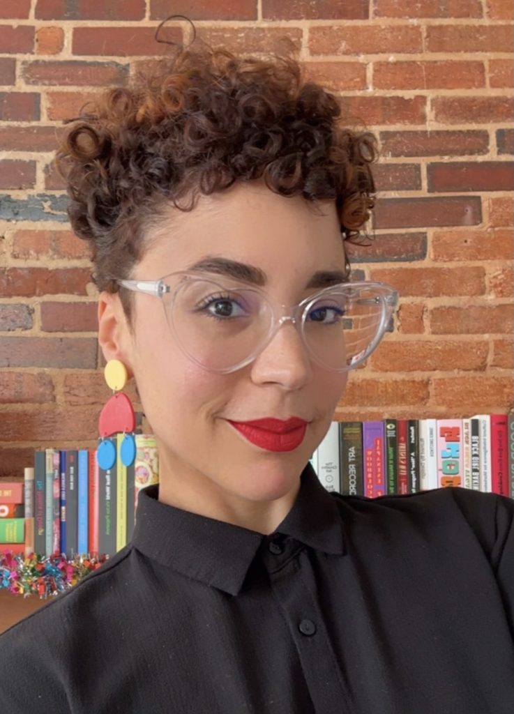 Photo of Natalia Arias in front of a brick wall and a low bookshelf.